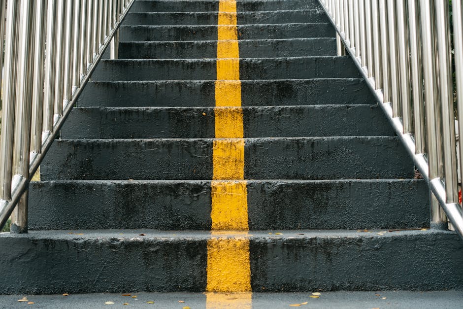 A metal outdoor staircase with anti-slip treads, featuring handrails on both sides, descends from a dark building structure to an exterior area. The stairs are constructed of steel with a diamond plat