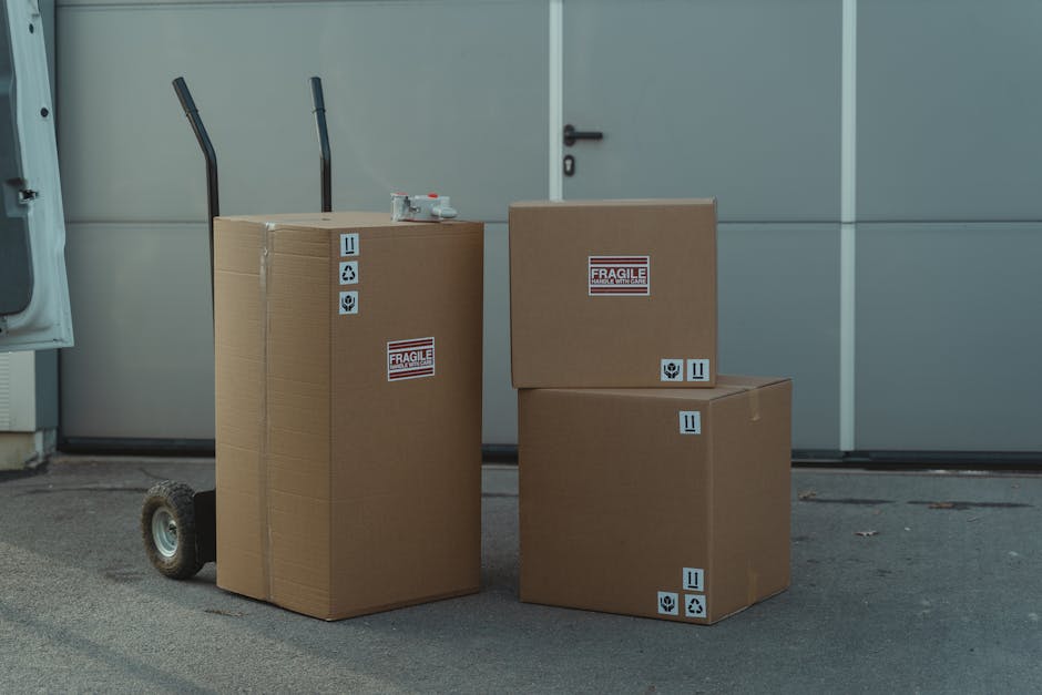 A person wearing a grey knitted sweater is seen lifting or preparing to carry a large cardboard box during a house relocation. The box is sealed with clear packing tape and features a red 'H' symbol,