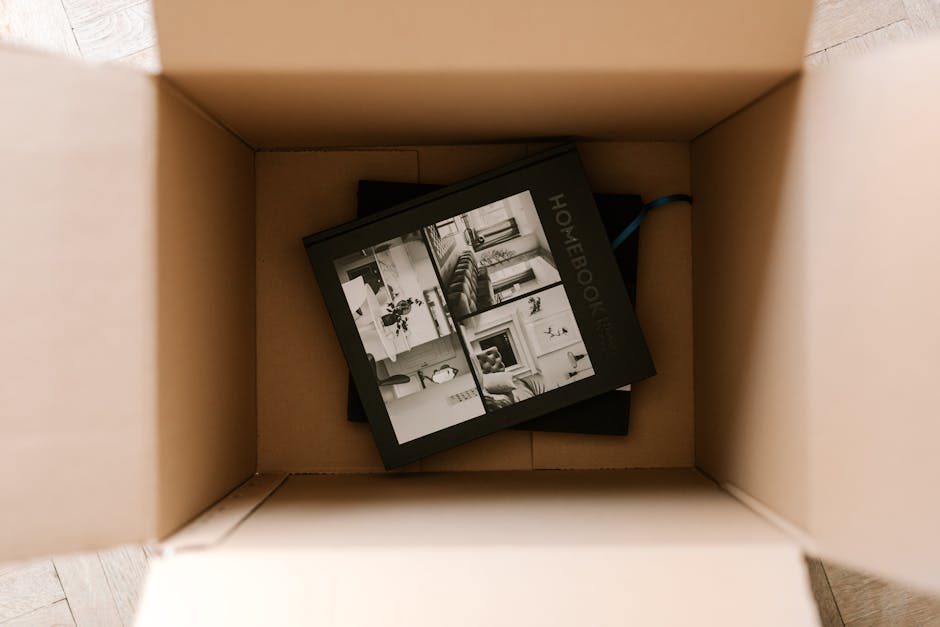 A young woman with long brown hair, dressed in a white tank top and beige pants, is sitting cross-legged on a wooden floor inside a room filled with several large cardboard moving boxes. She is smilin
