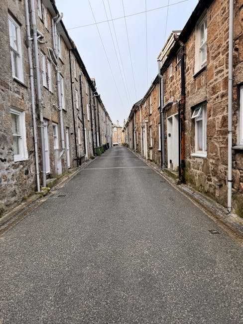 A narrow residential street with closely spaced terraced houses on both sides, constructed from brick and stone with white-framed windows. The pavement runs along the base of the buildings, and the ro