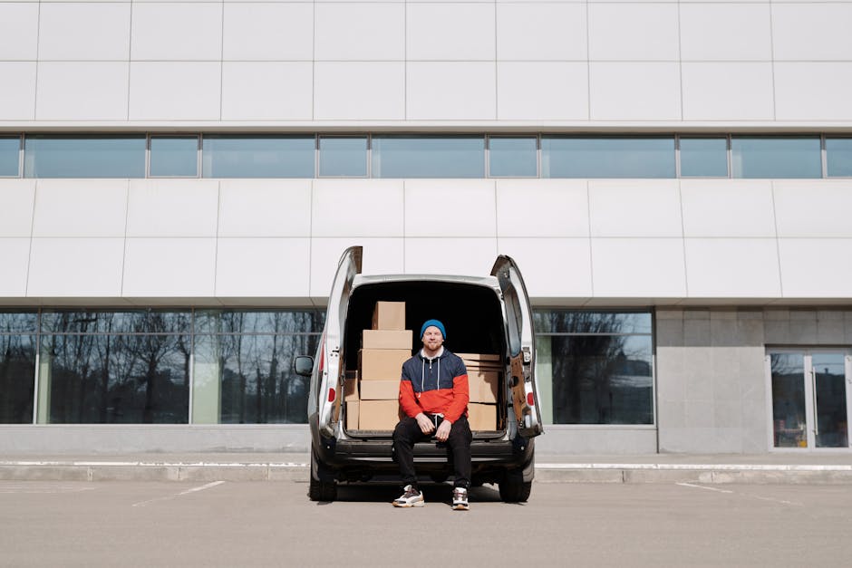 A young man wearing a white T-shirt and jeans is standing outside on the pavement next to a white moving van with its rear doors open, preparing boxes for loading. He is stacking cardboard boxes of va