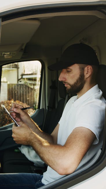 A man with a beard and wearing a black cap and white t-shirt is seated inside the driver's cab of a moving van, looking at his smartphone. The interior of the van shows a grey ceiling and part of the