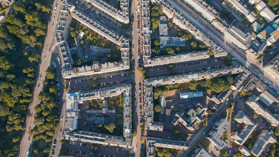 Aerial view of a residential area with tightly packed terraced houses and apartment buildings, situated along a network of streets and avenues. Several cars are parked along the roads and in designated parking areas, while trees and greenery are interspersed throughout the neighbourhood. The scene shows a typical urban environment with a mix of older and modern building structures, with some visible rooftops and pavements. The photograph captures the busy, organized layout of the streets within the residential district, with clear pathways for vehicle movement and pedestrian access. This image exemplifies a well-established urban community that may require professional house removals or moving services, such as those offered by Man and Van Holloway, to facilitate home relocation and furniture transport processes within the N7 postcode area.