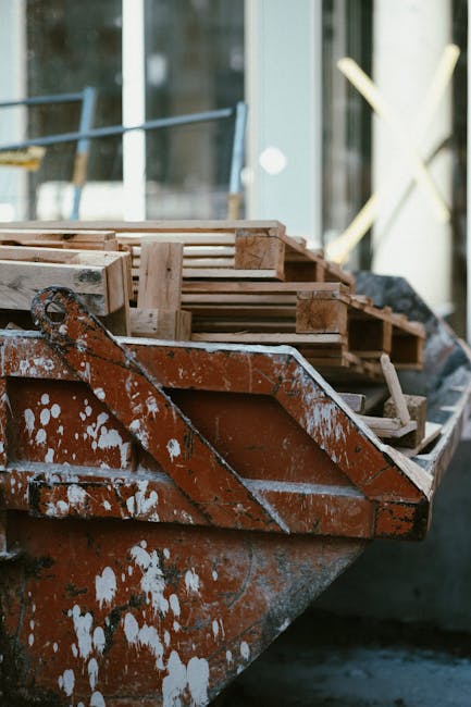 A close-up view of a rusty, orange-dyed workboat featuring chipped paint and visible white splatters, docked near a building with large glass windows. Inside the boat, wooden pallets and plastic conta