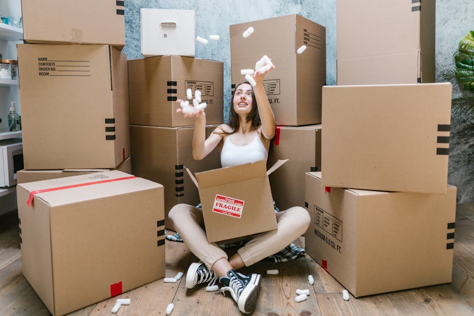 A woman sitting on the wooden floor inside a home surrounded by several large cardboard moving boxes, some with red and black tape and labels, with foam packing peanuts falling from her hands as she smiles. She is wearing casual clothing and sneakers, with an open box on her lap. The scene appears to capture a home relocation process, possibly during packing or unpacking stages. In the background, there is a textured wall, a small potted plant, and a kitchen countertop with a microwave and other items, indicating an indoor setting associated with house removals. The presence of multiple boxes and packing materials reflects a coordinated furniture transport or moving activity, typical of professional removals services such as those provided by Man and Van Holloway, experts in house removals and home relocation logistics.